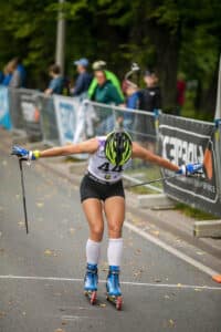 A professional roller skier crosses the finish line at the FIS World Cup on an asphalt track, wearing a helmet and competition gear, with spectators in the background.