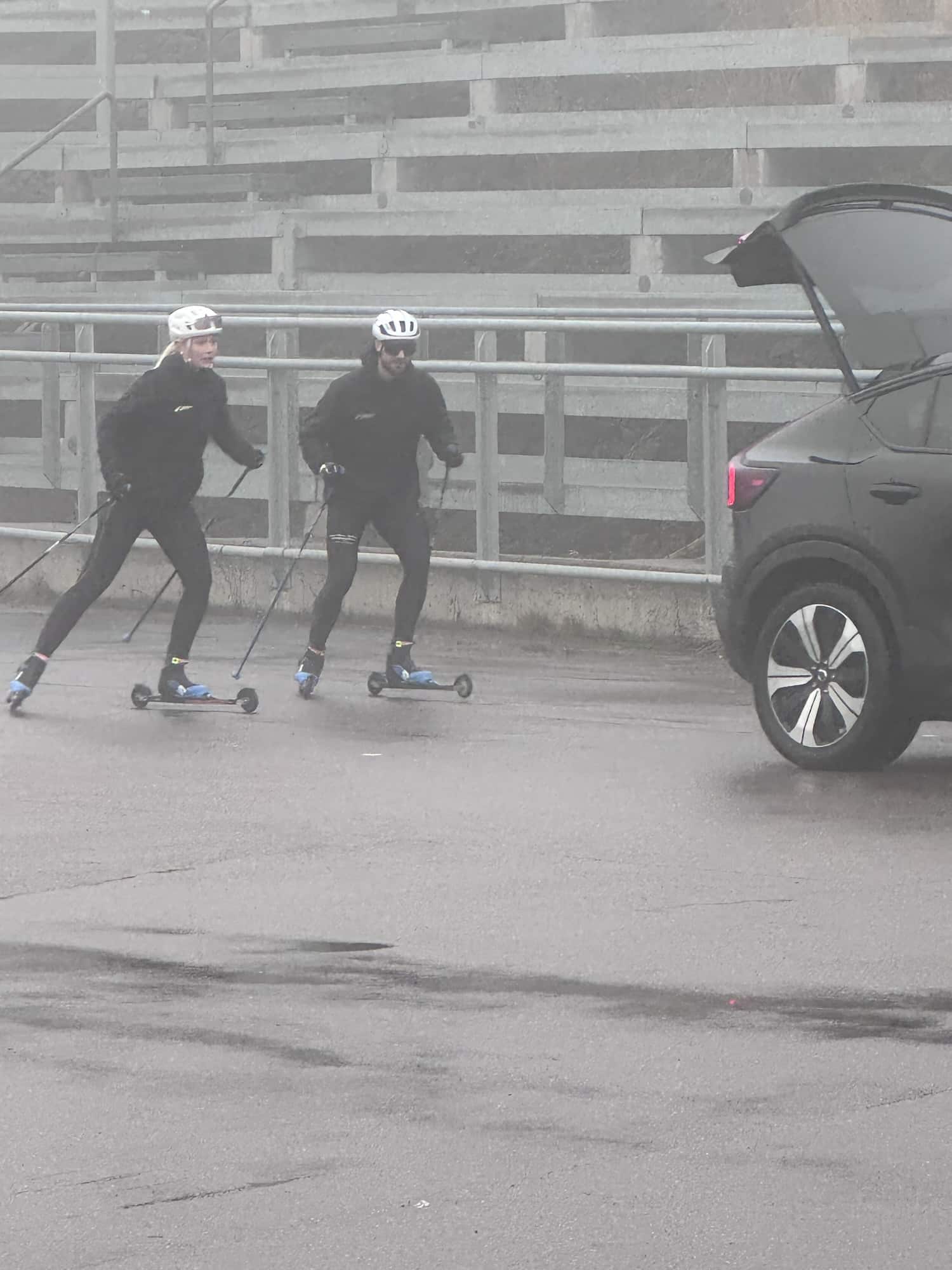 Two people skiing on roller skis in a foggy outdoor setting, wearing helmets and black athletic gear, with poles in hand. A car is parked in the background with its trunk open.