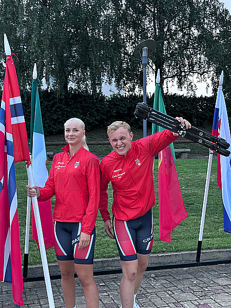 Two smiling FF Skis athletes display roller skis and the Norwegian flag at an outdoor event, ready for cross-country training.