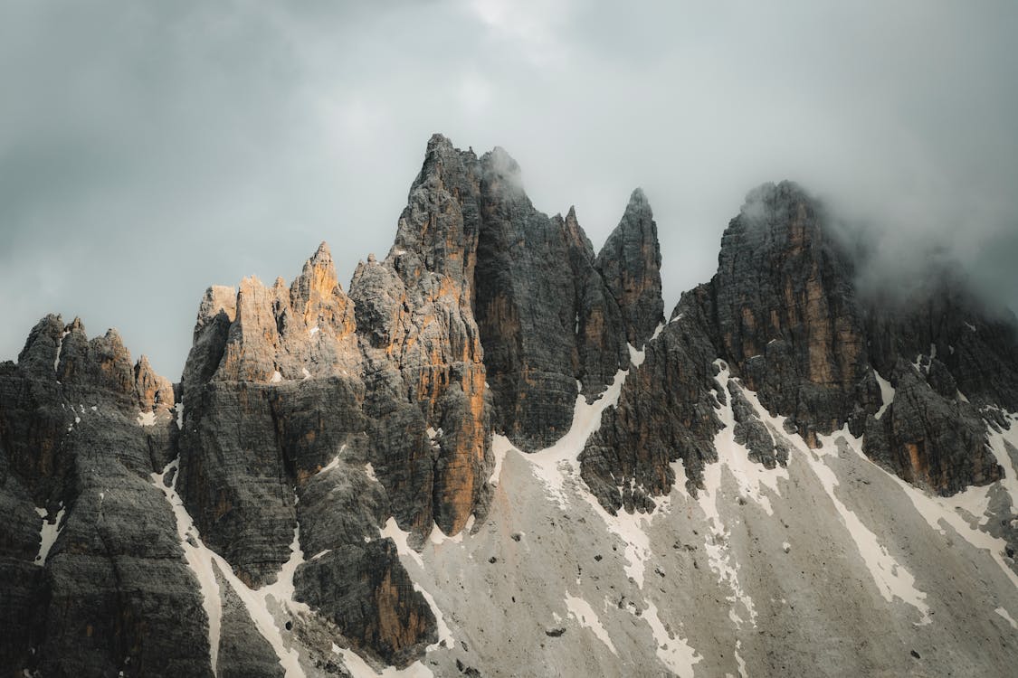 A close-up view of dramatic mountain peaks with jagged, rocky surfaces and patches of snow. Clouds linger around the top of the peaks, creating a misty and rugged atmosphere, with some parts of the mountains bathed in warm light from the sun.