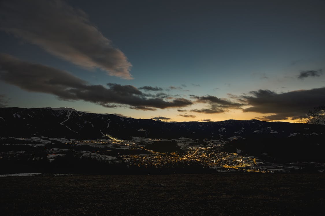 A panoramic view of a mountainous landscape at dusk, with a small town illuminated by streetlights at the valley's base. The sky has a blend of dark blue and orange hues as the sun sets behind the mountains, with some clouds scattered across the sky.