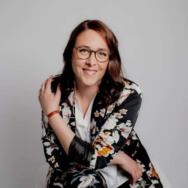 A woman with glasses wearing a floral patterned black blazer and a white shirt, smiling and resting one arm over her shoulder, sitting against a light gray background.